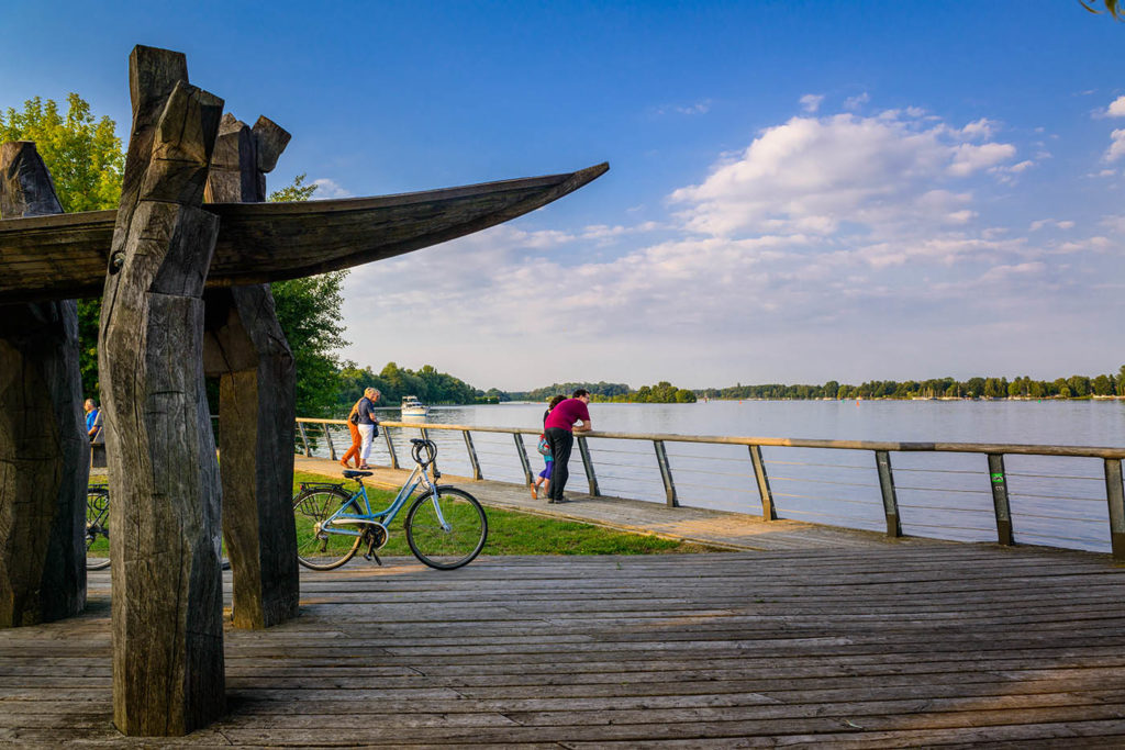 Uferpromende in Nieder Neuendorf. Menschen stehen am Ufer und schauen auf eine See. Blauer Himmel.