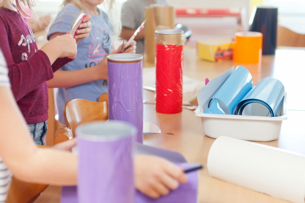 Kitakinder basteln. Es sind 3 Kinder zu sehen, die mit Scheren buntes Papier ausschneiden und Rollen bemalen. 